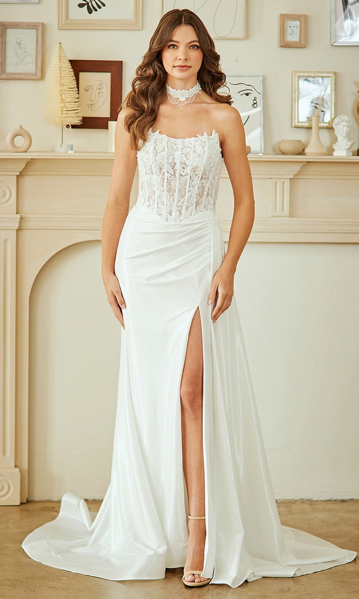 A woman poses indoors in front of a decorated mantel and neutral wall art, wearing the Amelia Couture 2075 Long Prom Dress in white—a floral lace corset gown with a high leg slit and matching choker.